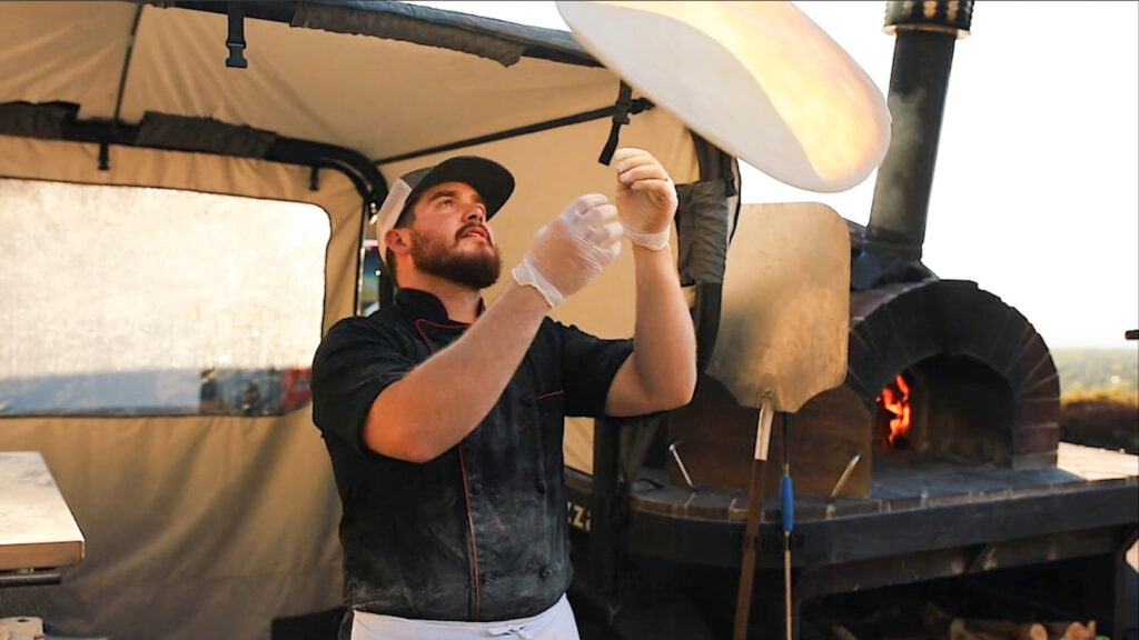 Italian chef throwing pizza dough in front of a wood-fired brick oven.